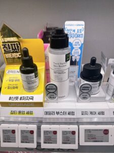 COSRX The 6 Peptide Skin Booster serum on the daily booster serum shelf at an Olive Young in Seoul, with the plus 76.4 percent skin texture panel and electronic price labels visible